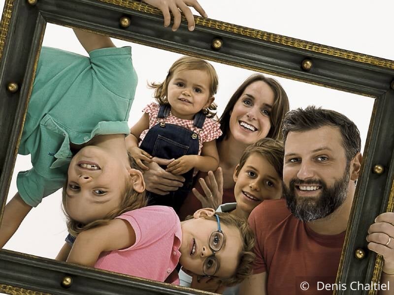 Photo de famille avec 4 enfants et 2 parents, ils tiennent un cadre et sourient.